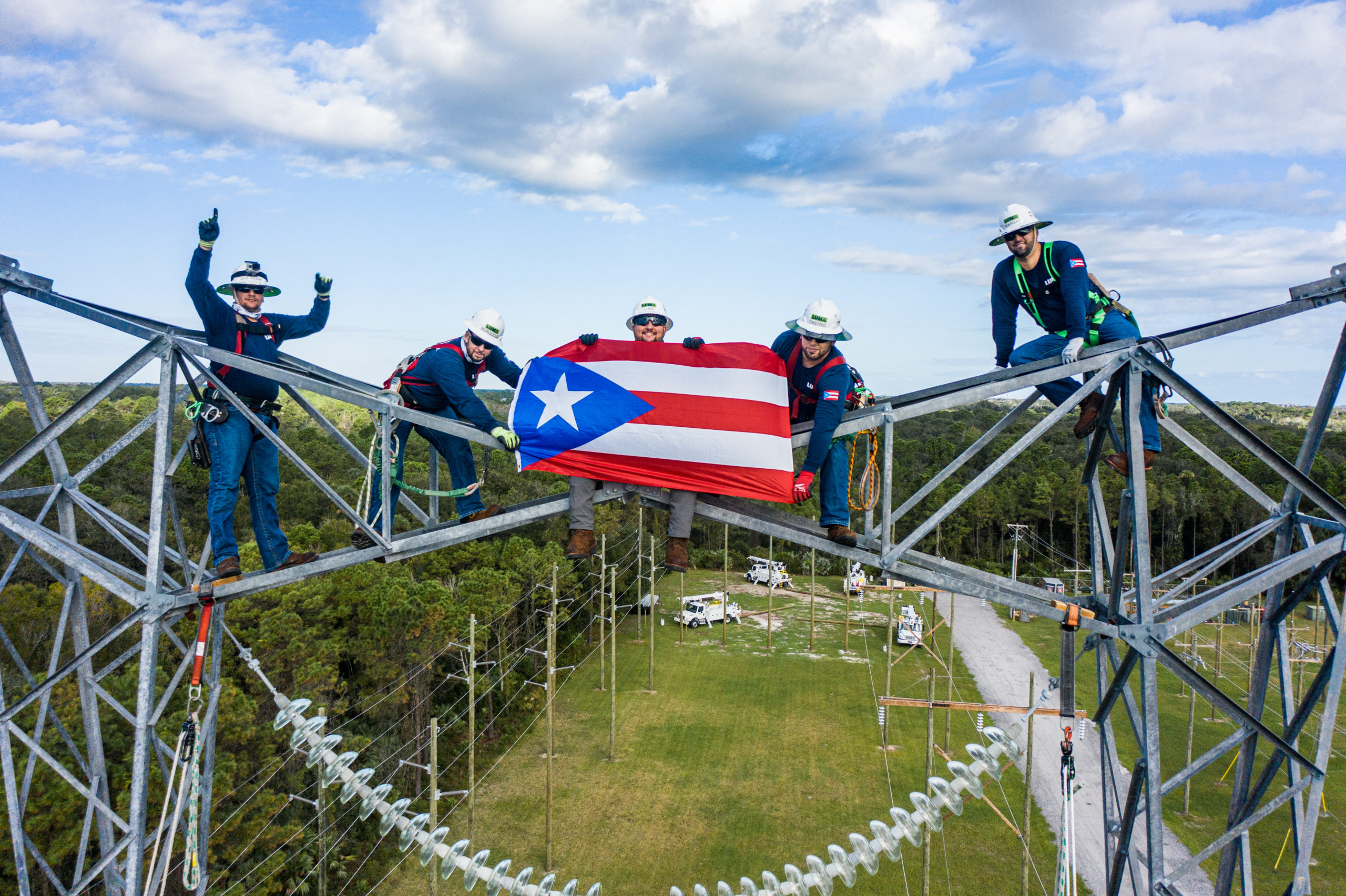 5 LUMA workers, smiling and holding the flag of Puerto Rico while hanging from an electric tower