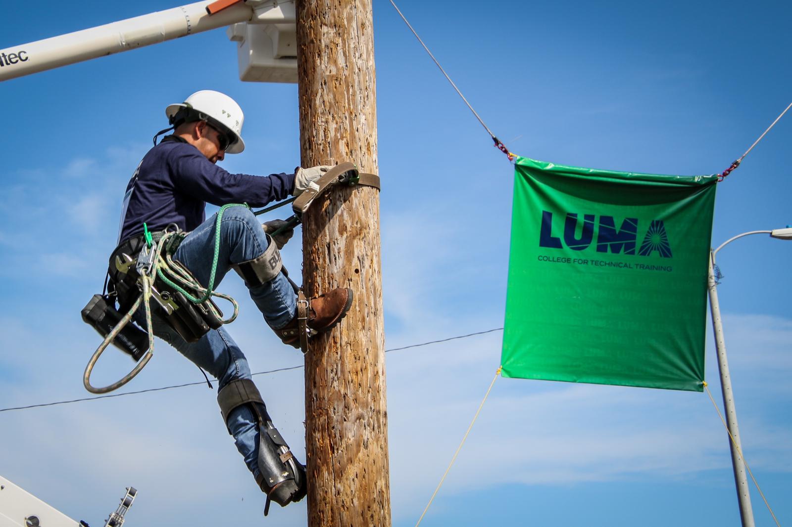 LUMA worker climbing on power pole using special equipment, next to a LUMA flag